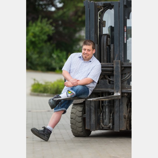 Kai Lehnert, ein Mann mit Behinderung, sitzt nach linksgebeugt auf der Gabel eines Gabelstaplers. Er tragt ein kurzärmeliges, fein, blau-weiß kariertes Hemd und eine kurze Jeanshose. An seinem linken Bein befindet sich eine Schiene. 